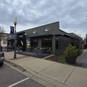 A modern dark green restaurant with outdoor seating on a cloudy day, located on a corner in a small town.