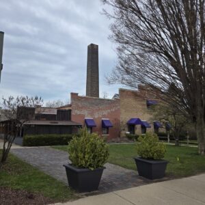 A brick building with purple awnings and a tall smokestack, surrounded by trees and planters on a cloudy day.