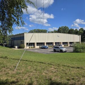 A beige industrial building with a parking lot and a few cars, surrounded by grass and trees under a blue sky.